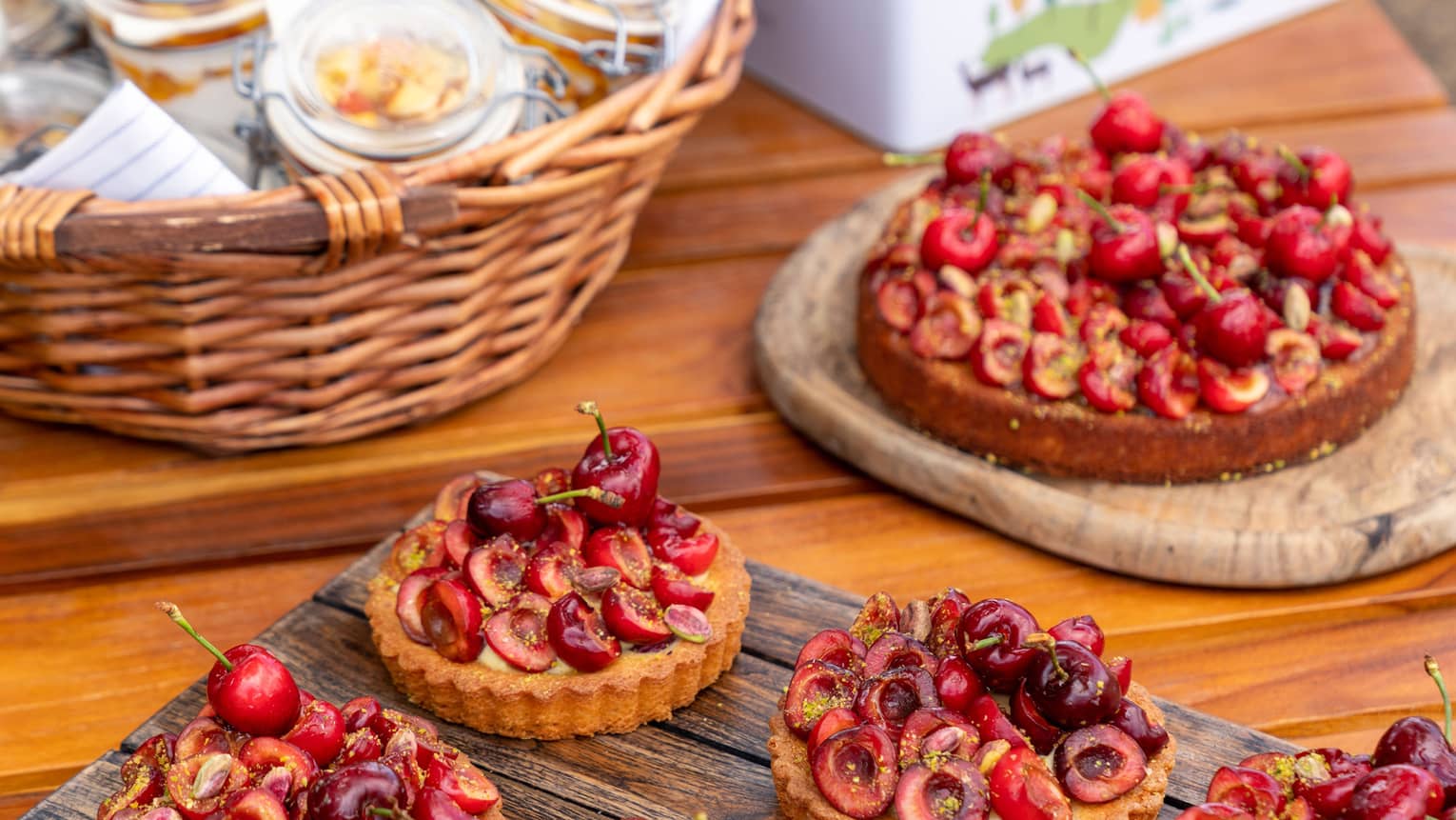 Artfully placed on a wooden table, a large cherry fruit flan with six small ones on rustic trays, and a basket of glass jars.