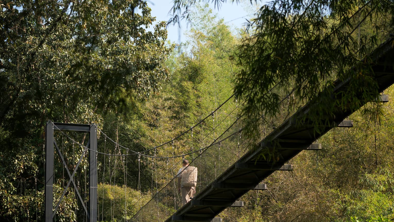 Man with woven basket on back walks across suspension bridge over tropical trees