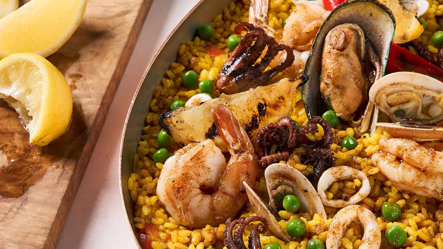 Seafood paella with assorted seafood in traditional oval-shaped metal pan, lemon wedges on cutting block