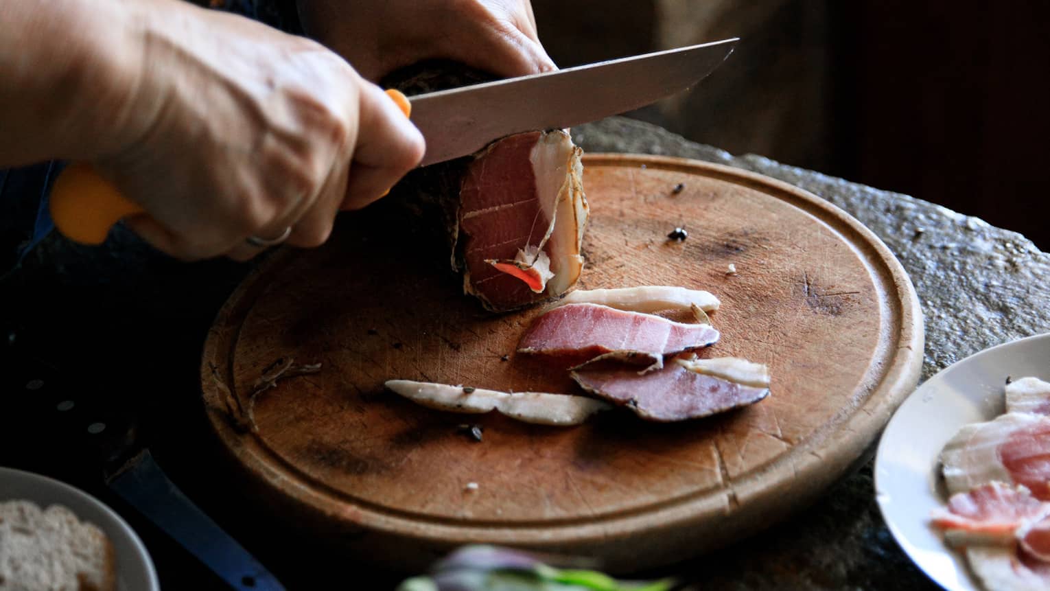 Person cutting Louza (Aegean prosciutto) on round cutting board