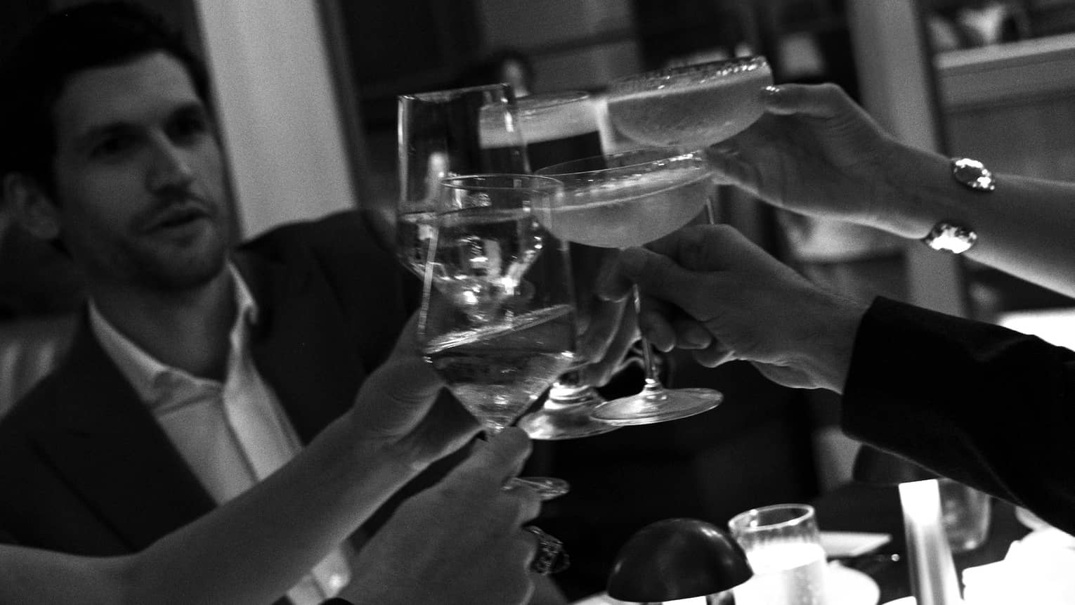 Black and white photo of friends raising their wine and cocktail glasses to cheers around a dimly lit restaurant table