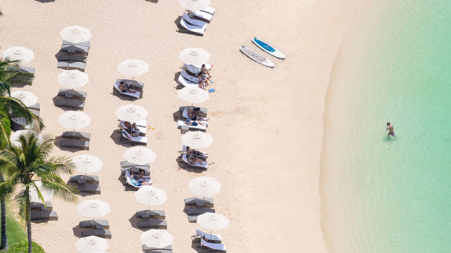 Aerial view of person walking out of water onto beach sand, lounge chairs and umbrellas