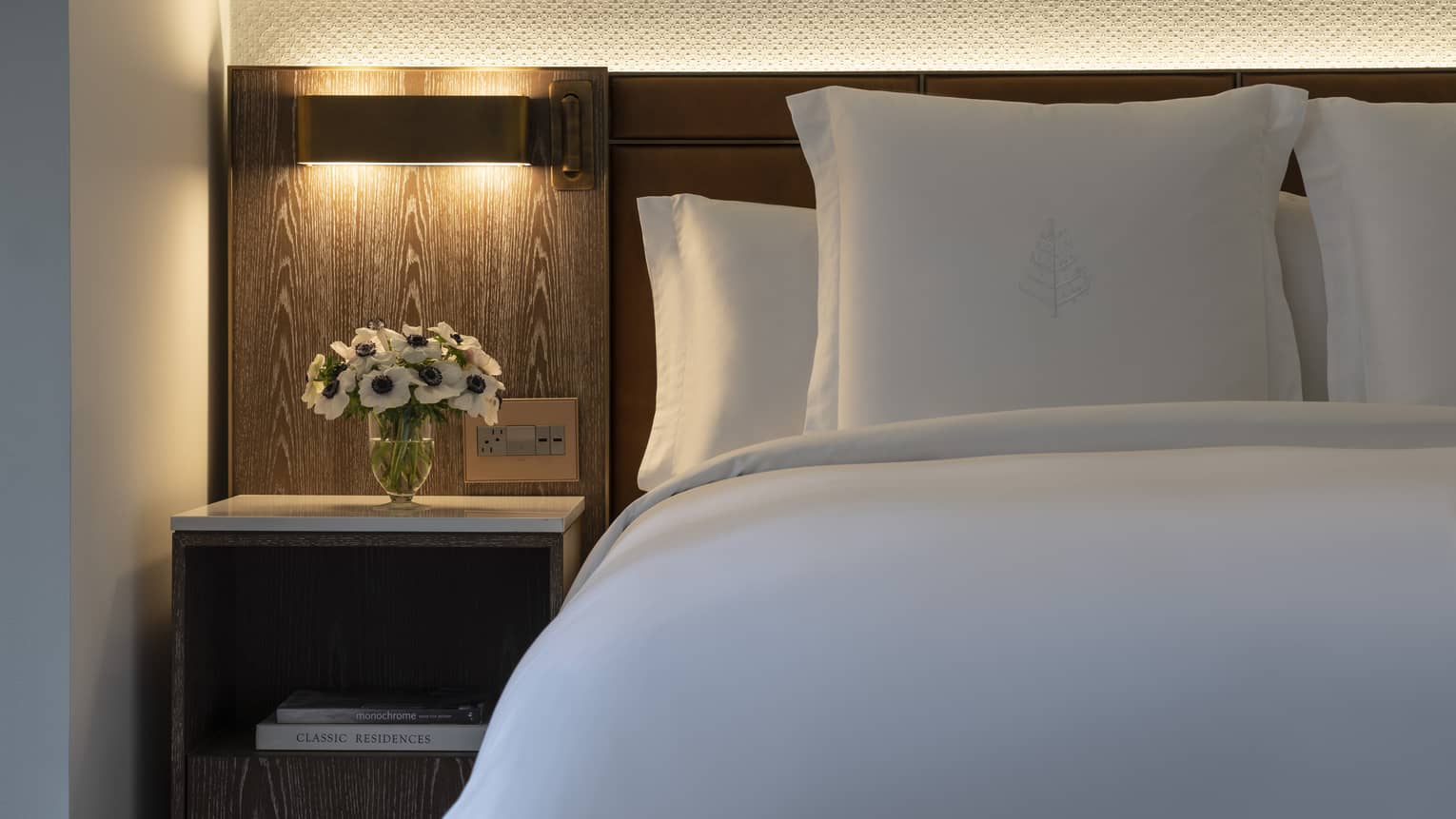 Close-up of bed with white linens and pillows, light above bedside table with vase of flowers