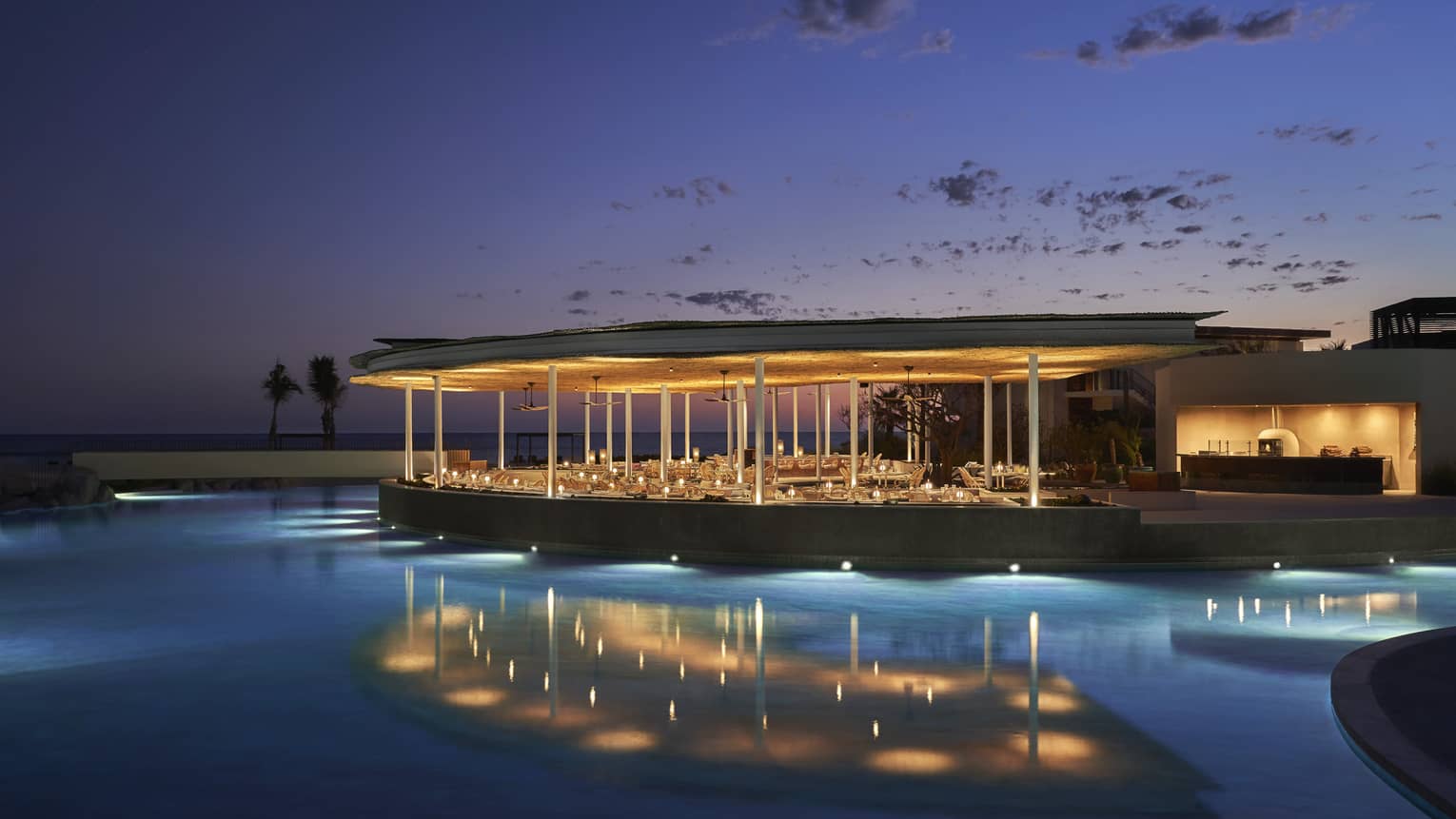 The exterior of an outdoor eating area at night on the water of a shore, it is well lit with lights.