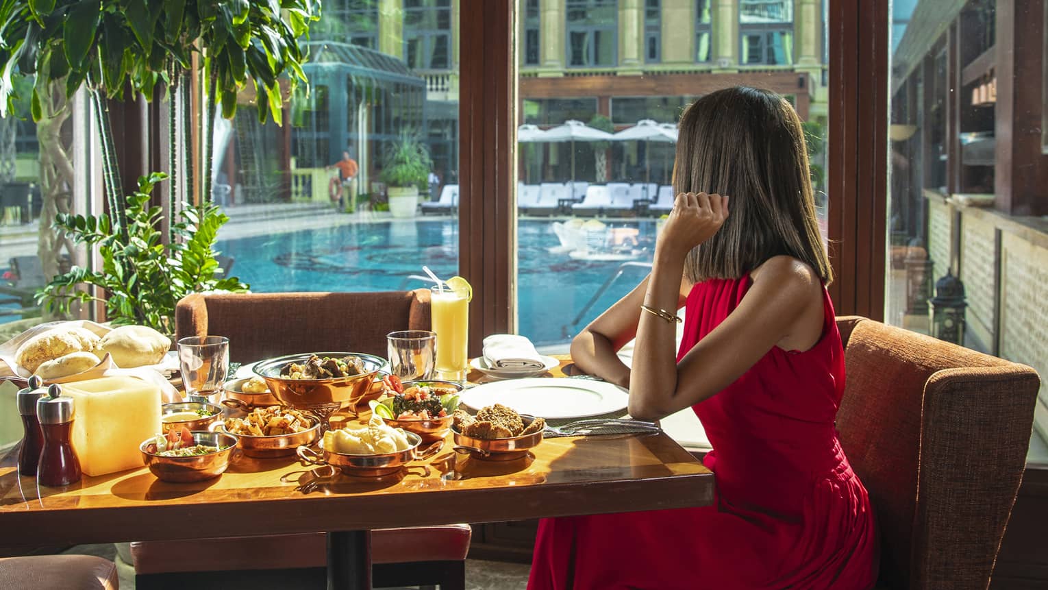A woman in a long red dress sits at a table with a variety of food next to a long window overlooking a pool