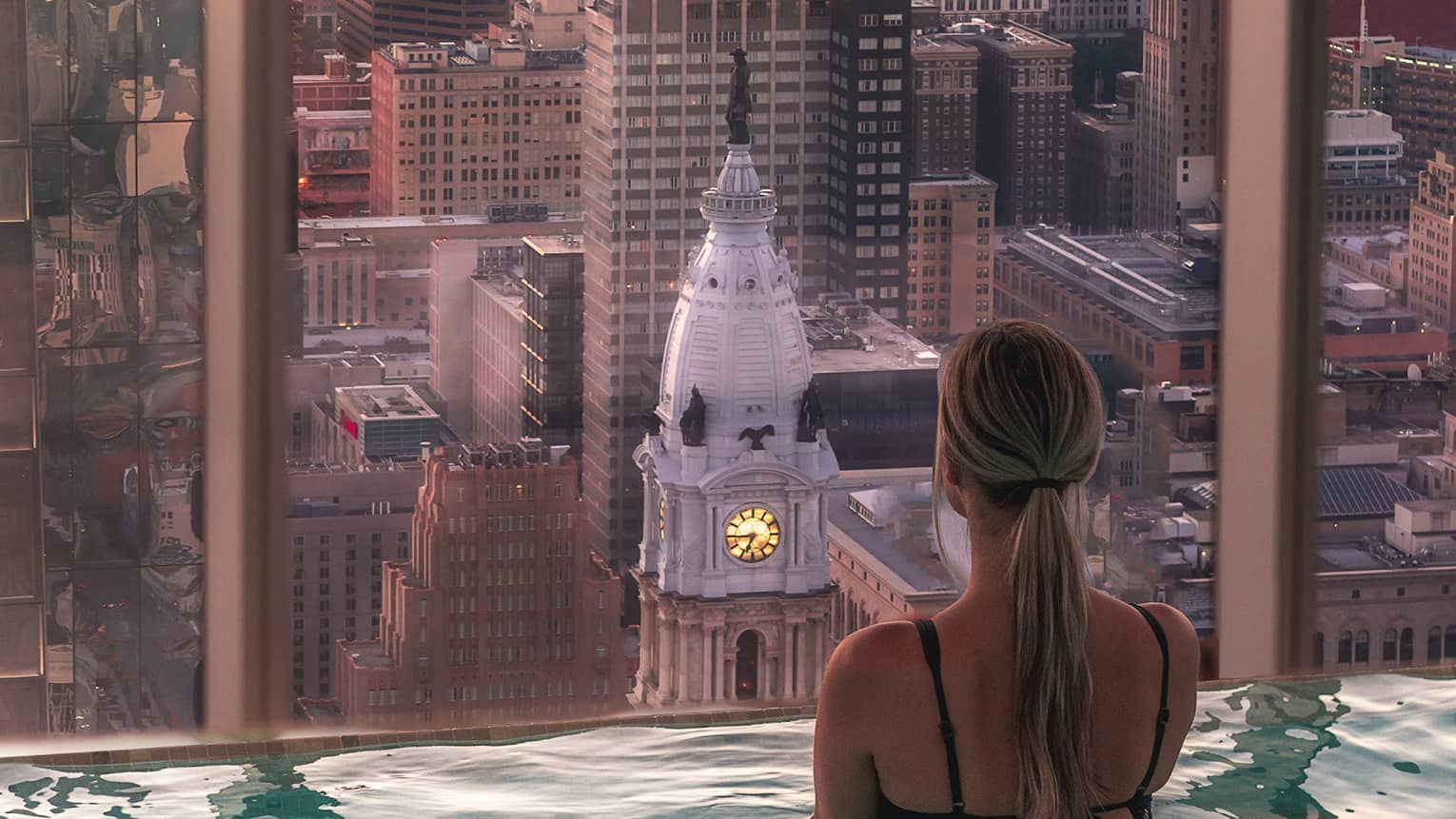 A woman standing in a pool in a tall building looking out a large window at a city