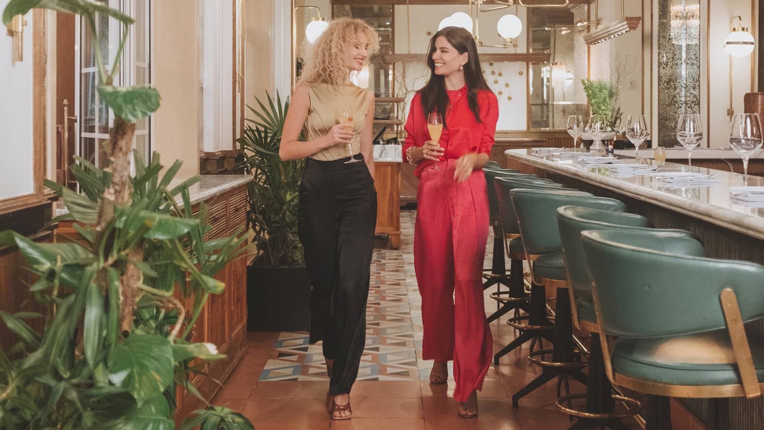 Two women walking a well lit restaurant with colourful tiles and seat at a bar.