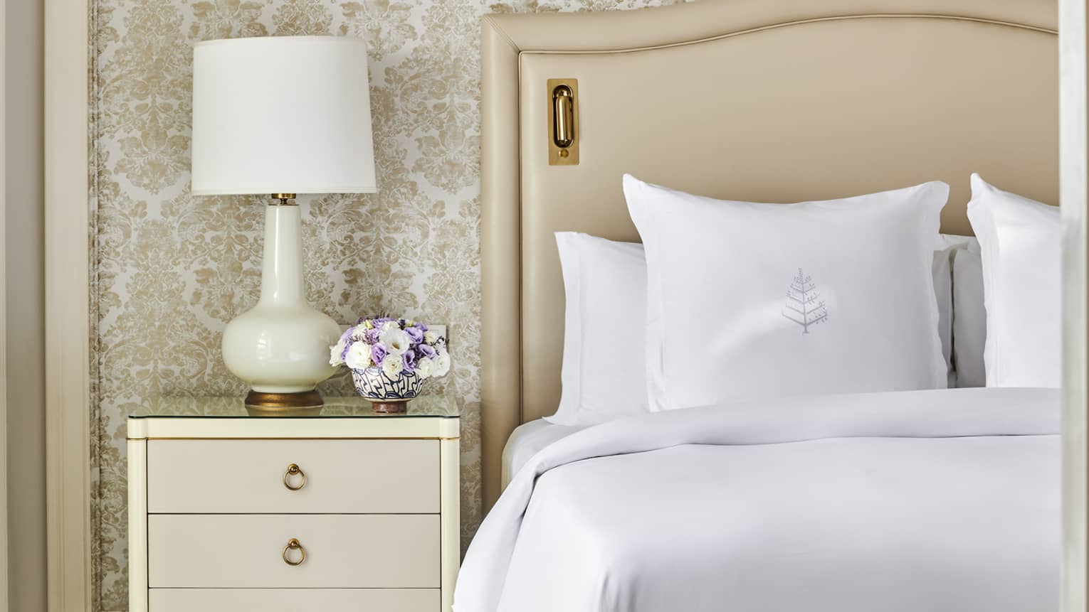 Guest bedroom with king bed, white linens and cream tufted headboard, foot bench, three-drawer side table with lamp, paisley wallpaper