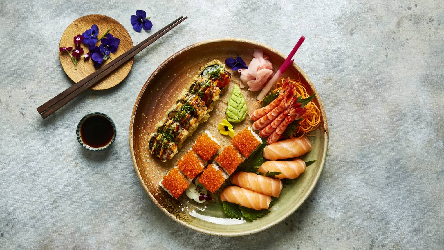 A ceramic bowl holding sushi and sashimi rolls, a small container of soy sauce, a small ceramic plate holding chopsticks and small purple flowers