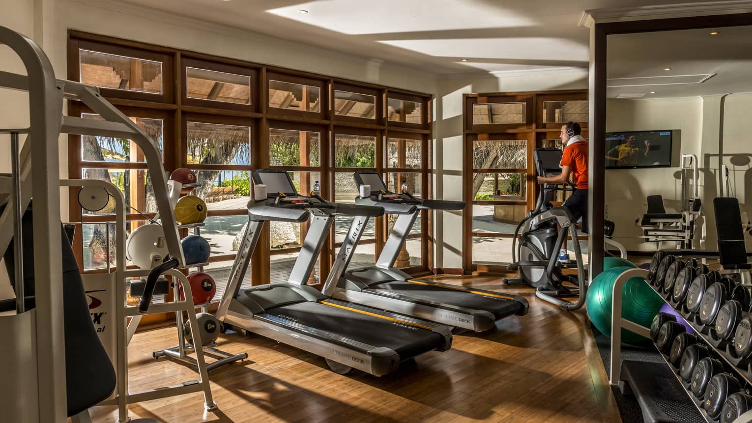 Gym with wooden floors, two treadmills, person on elliptical, free wights, windows looking out to beach