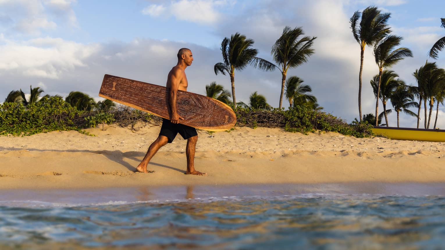 Surfboard designer Bonga Perkins walks along the water's edge with intricately carved wooden surfboard, palm trees swaying in the distance