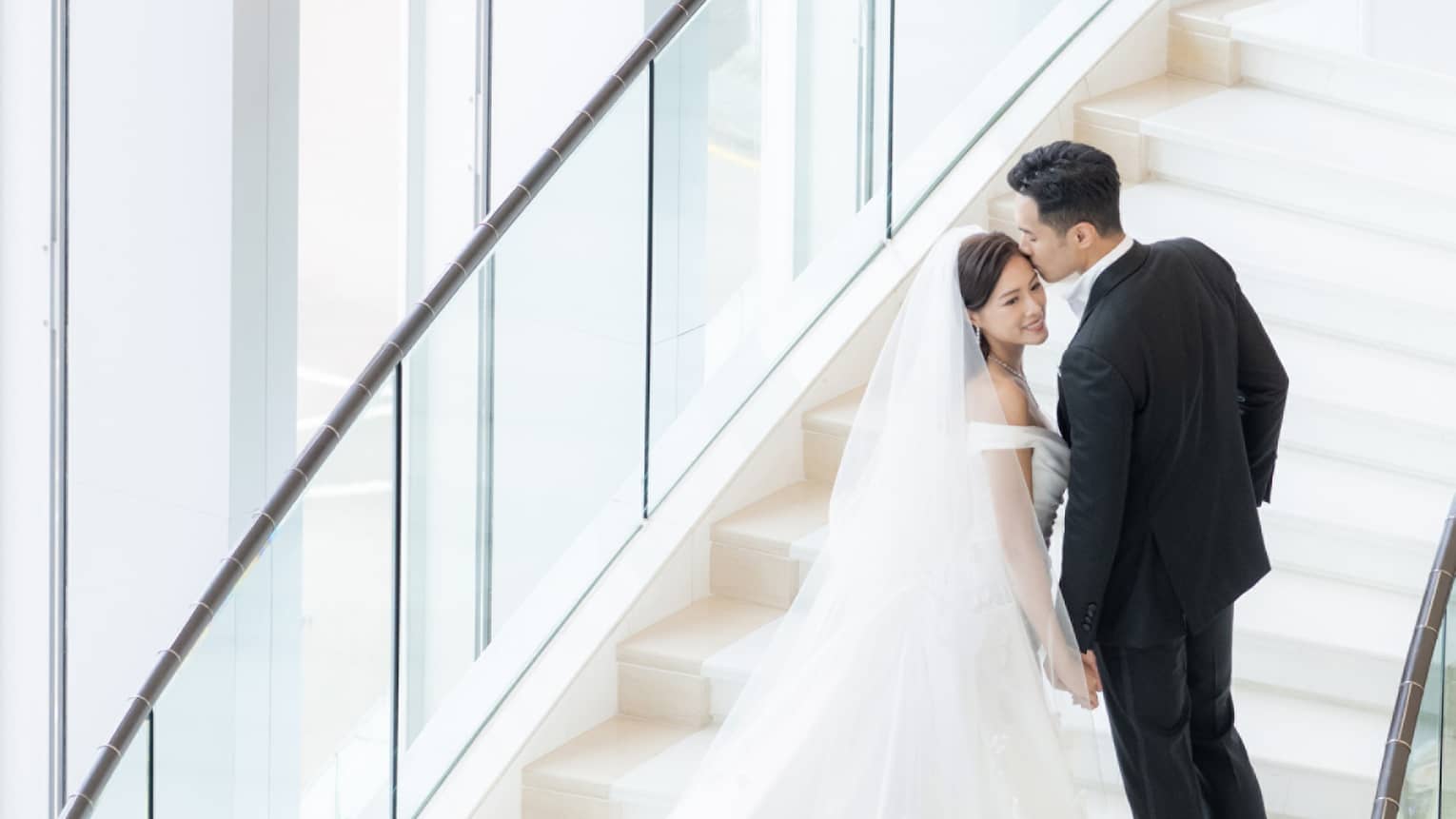 A bride and groom walking up stairs near large windows.