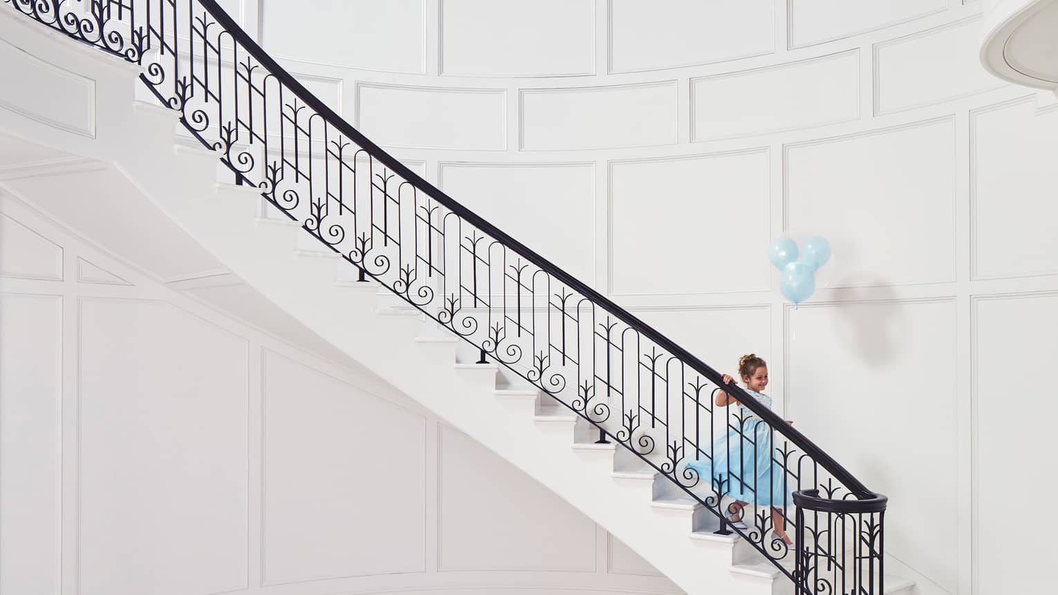 Child wearing blue gown with blue balloons walks down elegant white staircase with black iron railing