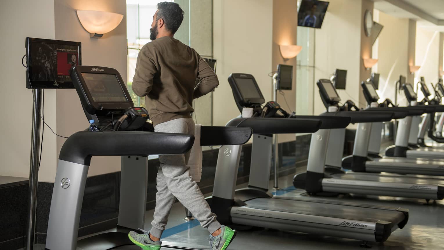 Back of man in sweatshirt and sweatpants as he runs on treadmill in gym next to row of cardio machines