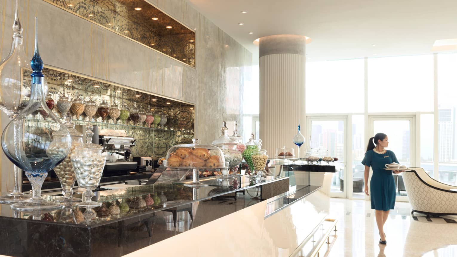 Server with tray walks through sunny Al Meylas Lounge near bakery counter with glass vases 