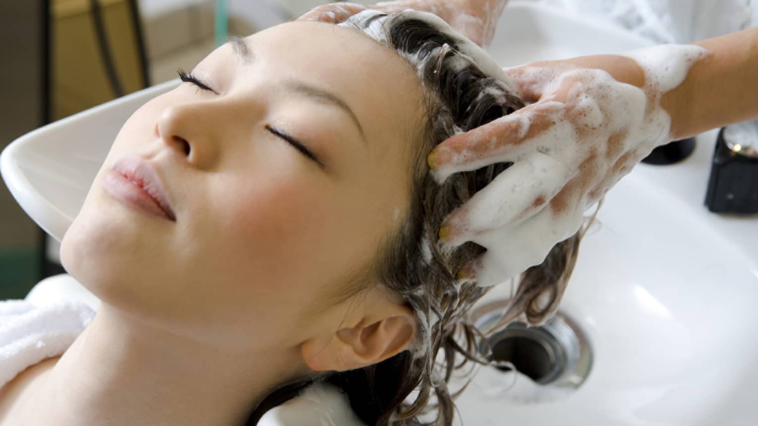 Woman closes eyes and leans back against sink as hands massage shampoo into her scalp