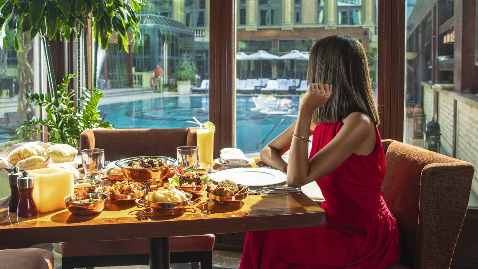 A woman in a long red dress sits at a table with a variety of food next to a long window overlooking a pool
