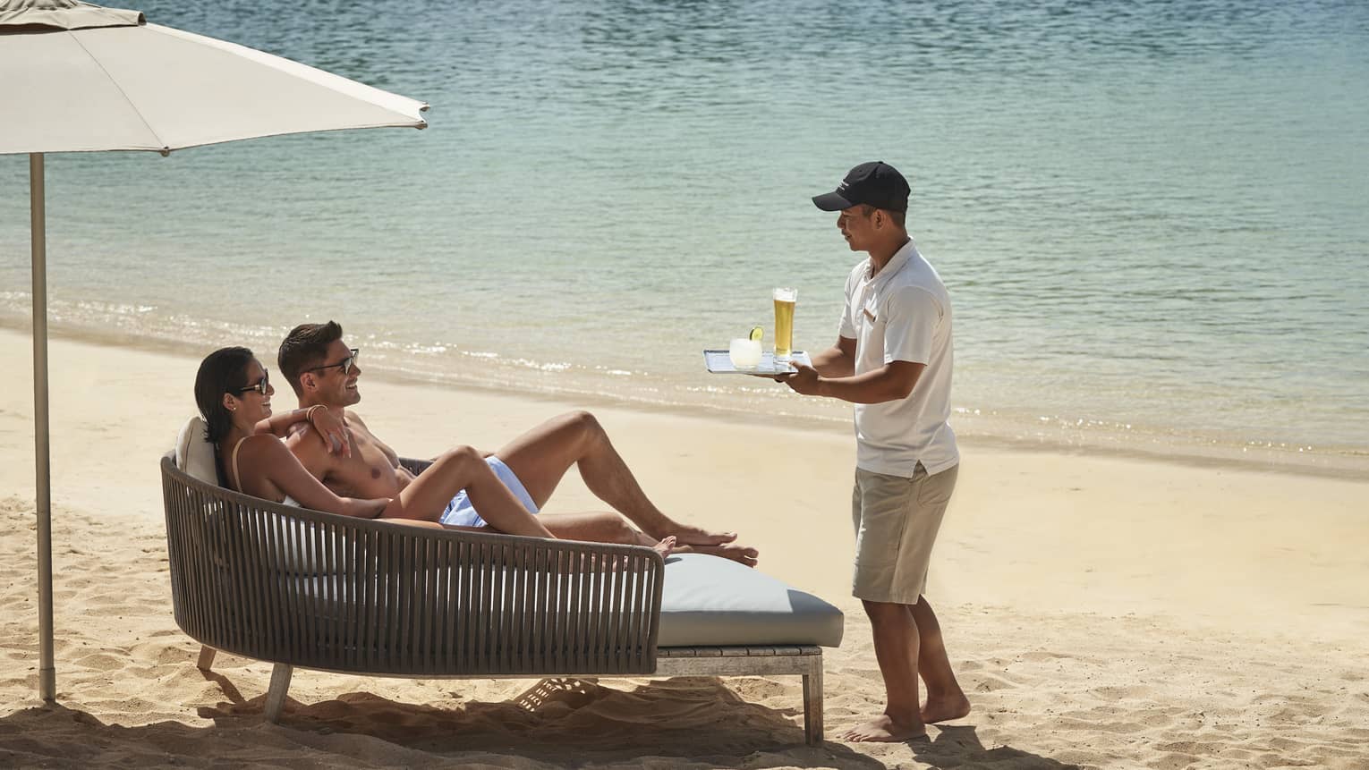 A couple lounges on a daybed on the beach while a beach attendant brings them cocktails on a tray