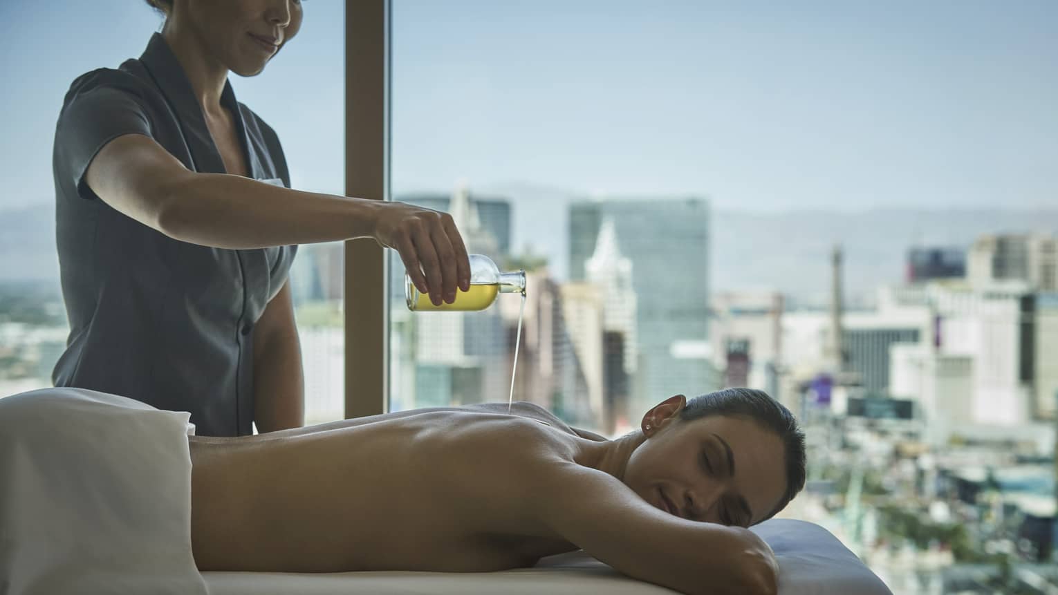 A woman with eyes closed lies on massage table and receives a treatment in a room with a view of the city skyline