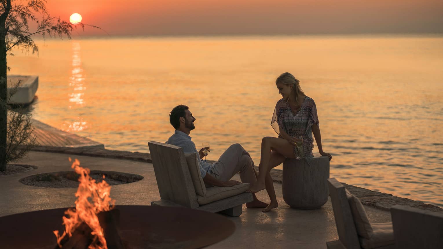 Couple lounges by Taverna Fire Pit on modern patio overlooking ocean at sunset