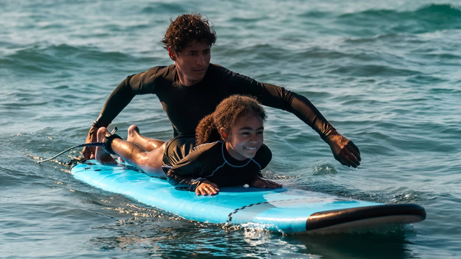 Male suf instructor wearing a black long-sleeve rashguard helps a young surfer paddle in the ocean
