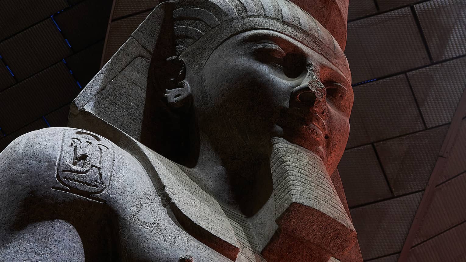 Close-up on a dark stone statue of a bearded pharoah with three heiroglyphic carvings on his chest and shoulders.