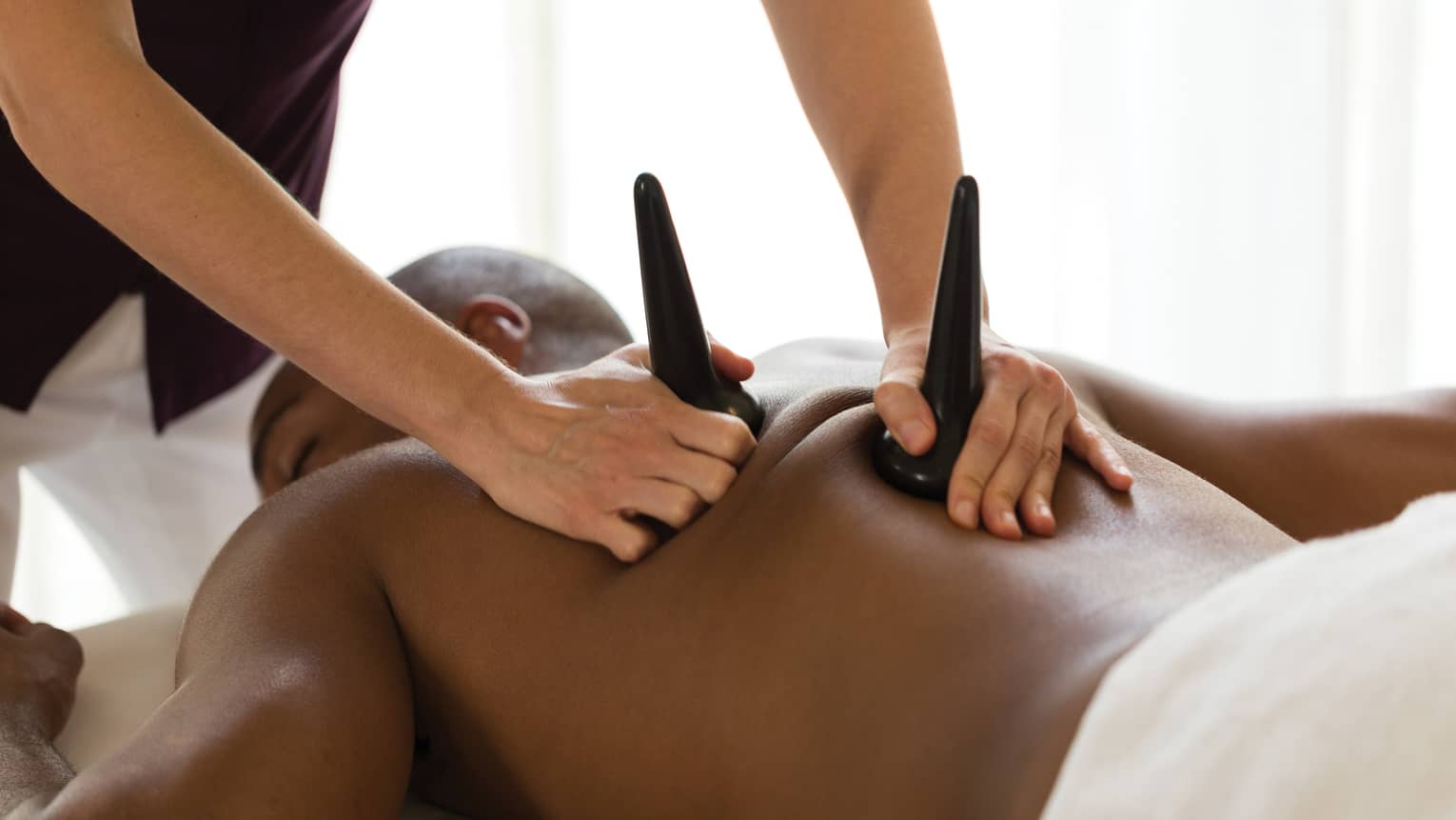 Masseuse pressed sculpted basalt stones on man's bare shoulders during massage