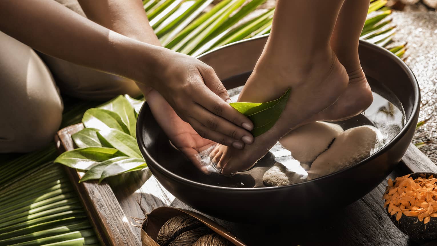 Close-up of woman's feet in round wood bowl as hands run tropical leaf over foot