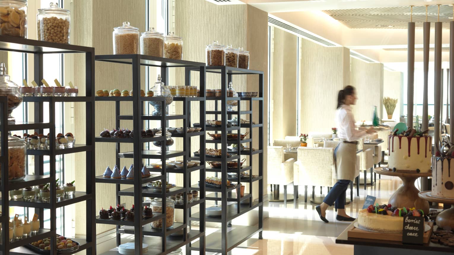 Three tall racks filled with fancy pastries and desserts in front of window, server carries tray to dining room