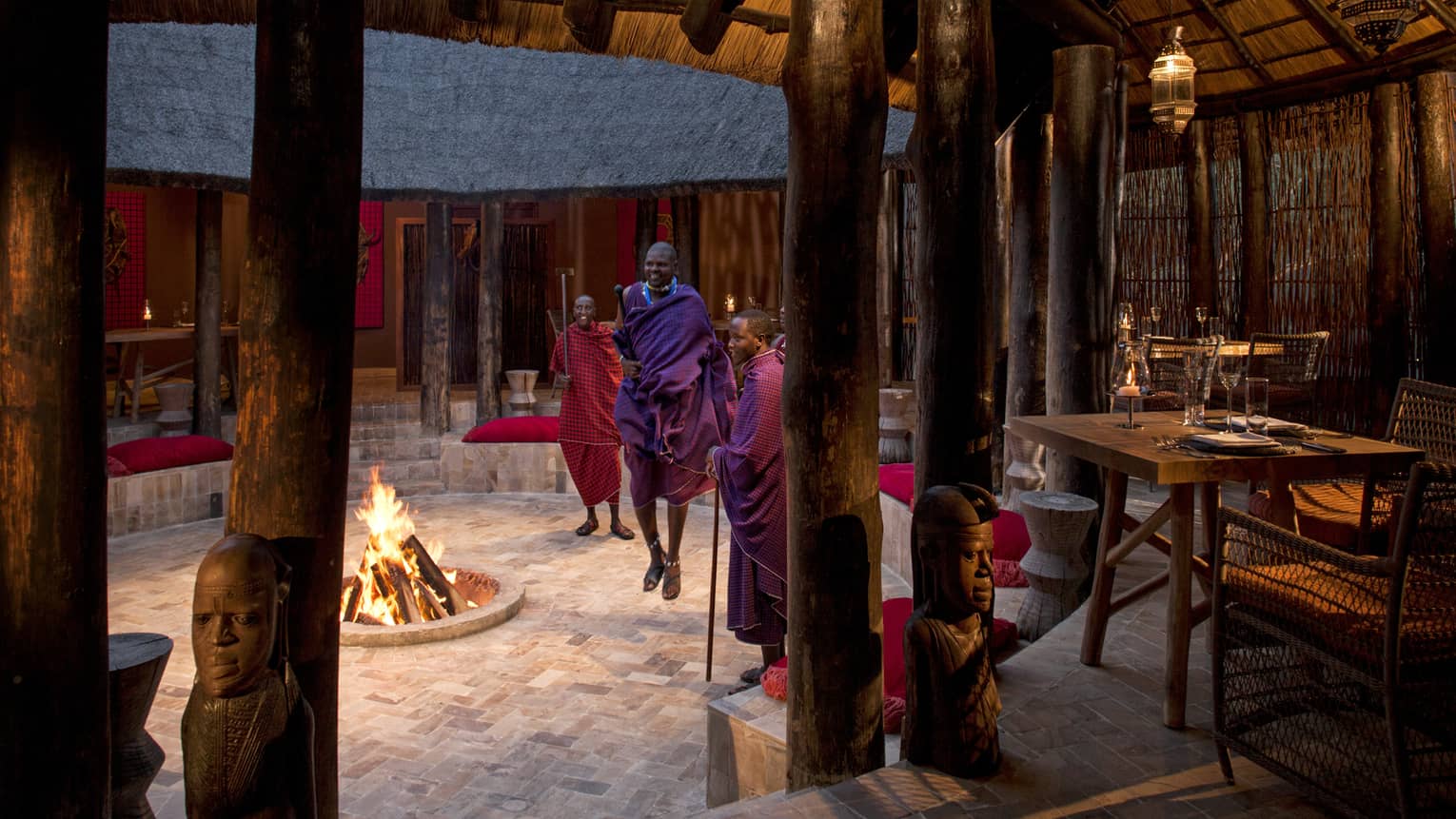 Maasai perform around a fireplace under a thatched hut with rustic tables and chairs at Boma Grill