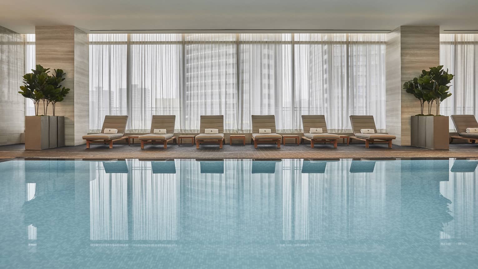 A wide angle view of the Four Seasons Toronto's indoor pool lined with cream lounge chairs and indoor potted trees. 