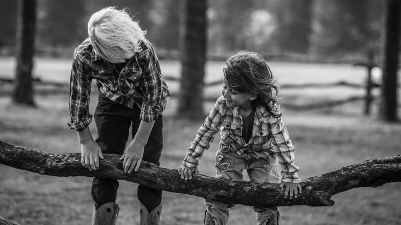 Black and white shot of two kids in cowboy boots leaning over the railing of a log fence