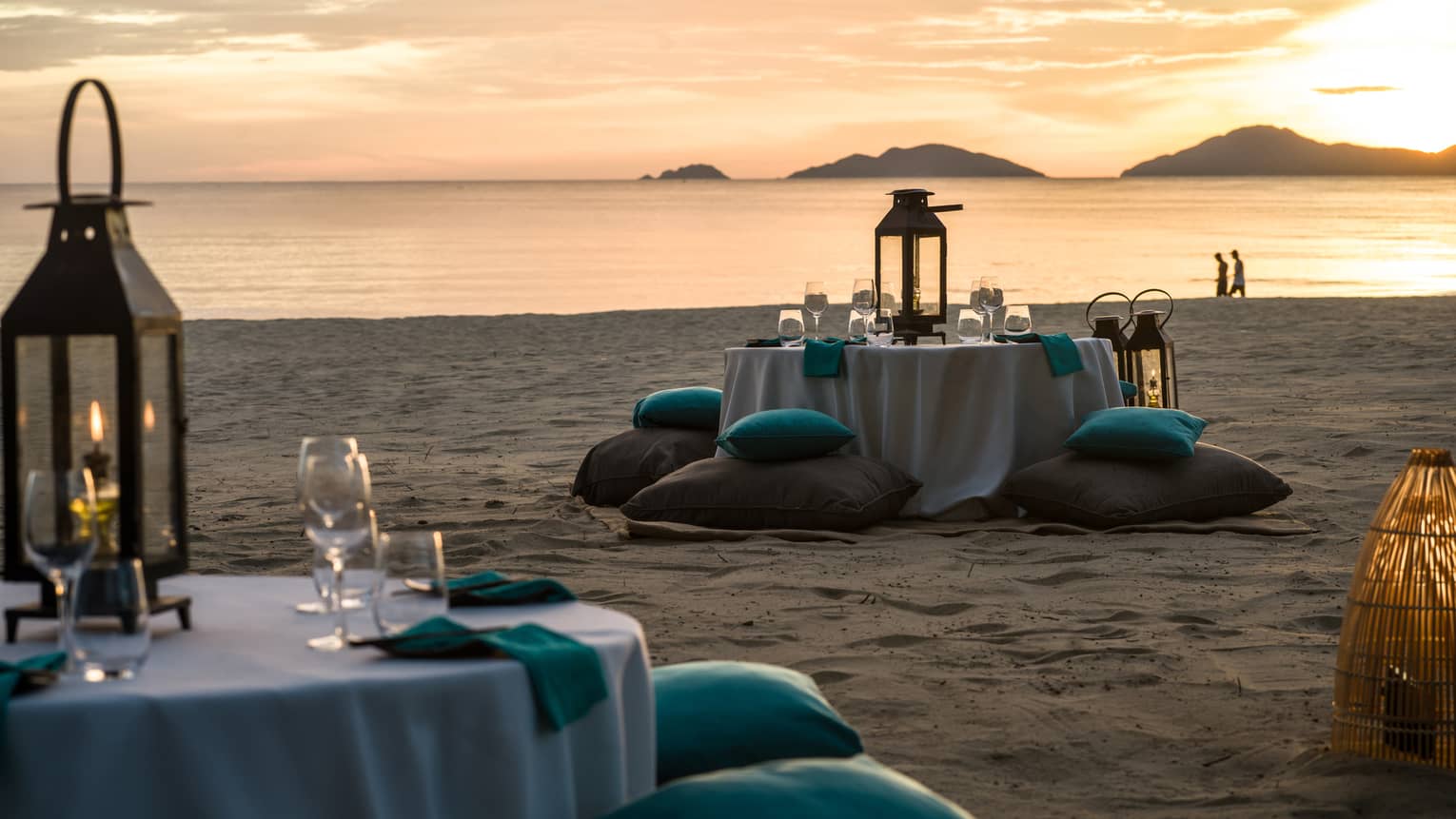 Two tables with white cloths, glasses, lanterns and turquoise cushions on sandy beach at dusk 