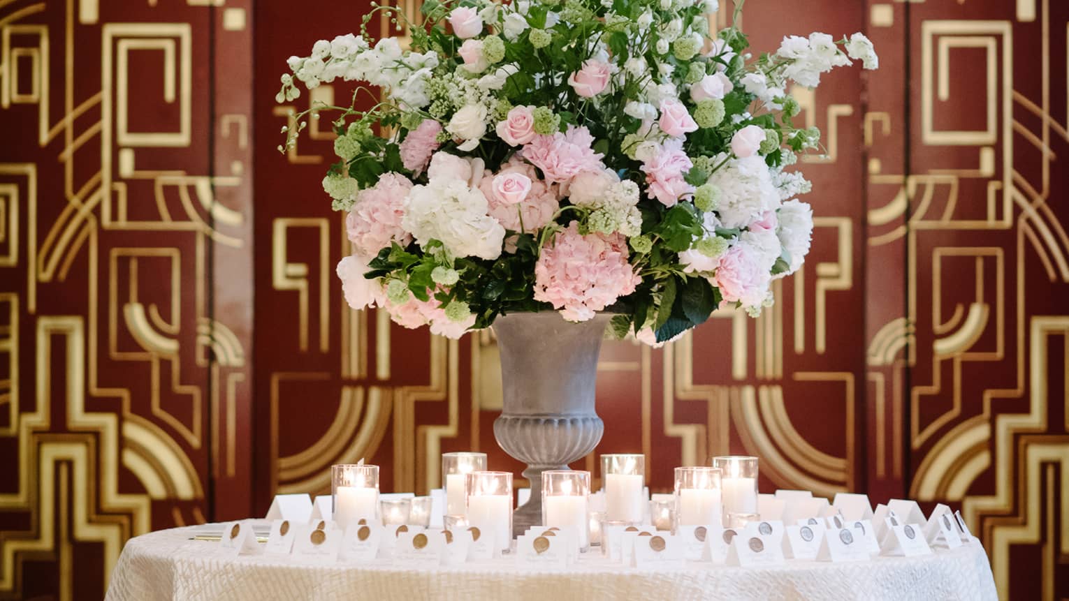 Glowing candles and name cards around vase with pink and white flowers against Art Deco wall