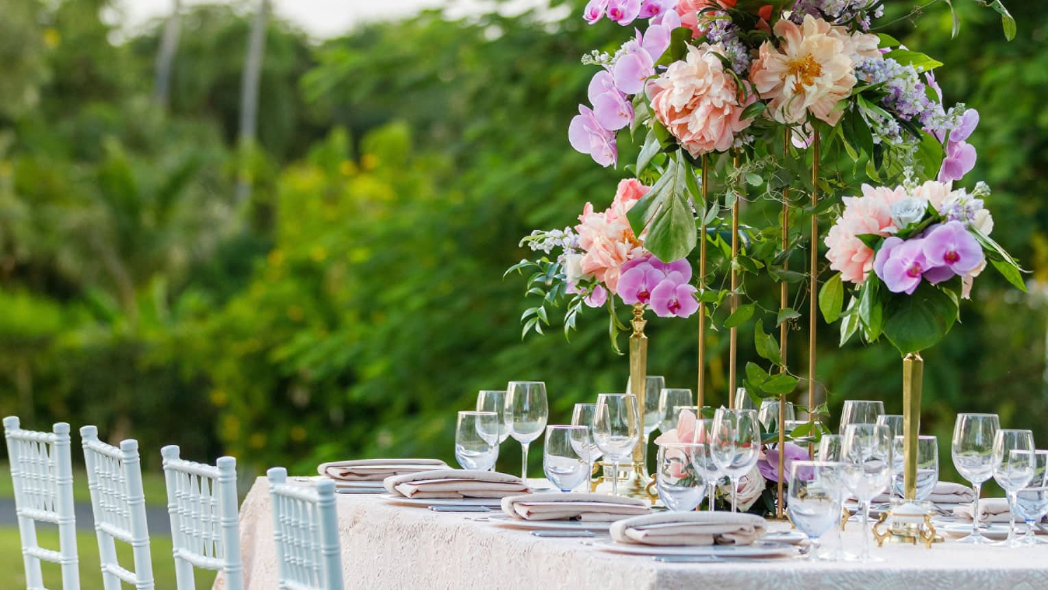 Angled set table, pink tablecloth, four white chairs, colourful floral centrepiece, trees in backdrop