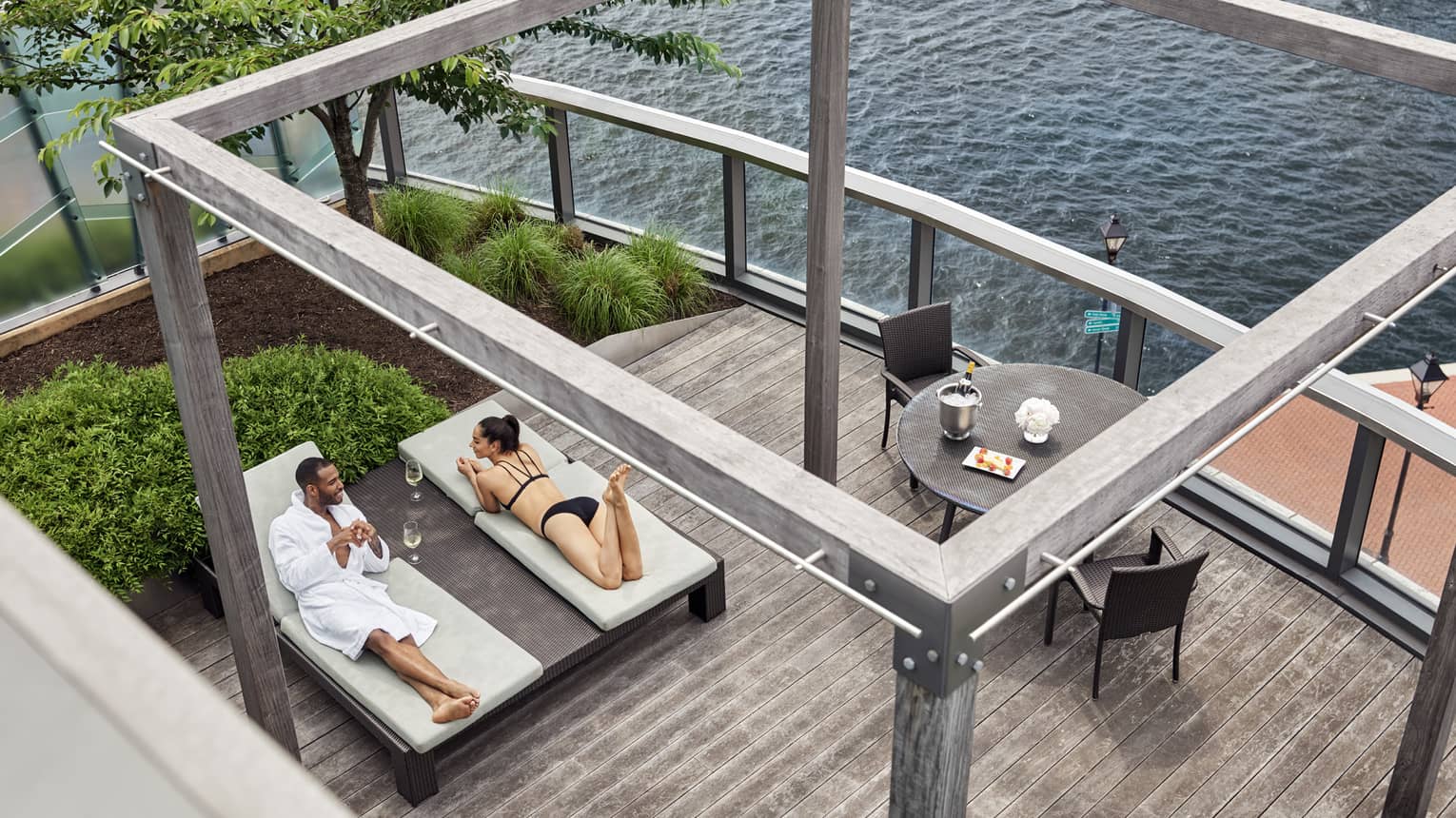 Couple laying on lounge chairs on wooden outdoor decking, table and chairs beside water