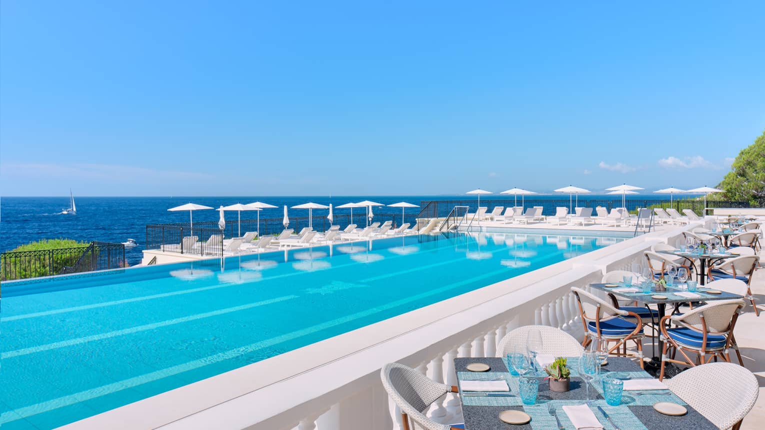 Mediterranean seafront infinity pool surrounded by white tables, lounge chairs, and umbrellas