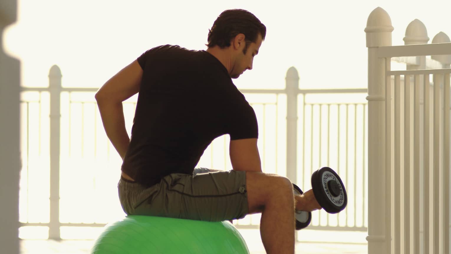 Back view of man in workout gear with hand weight, seated on exercise ball