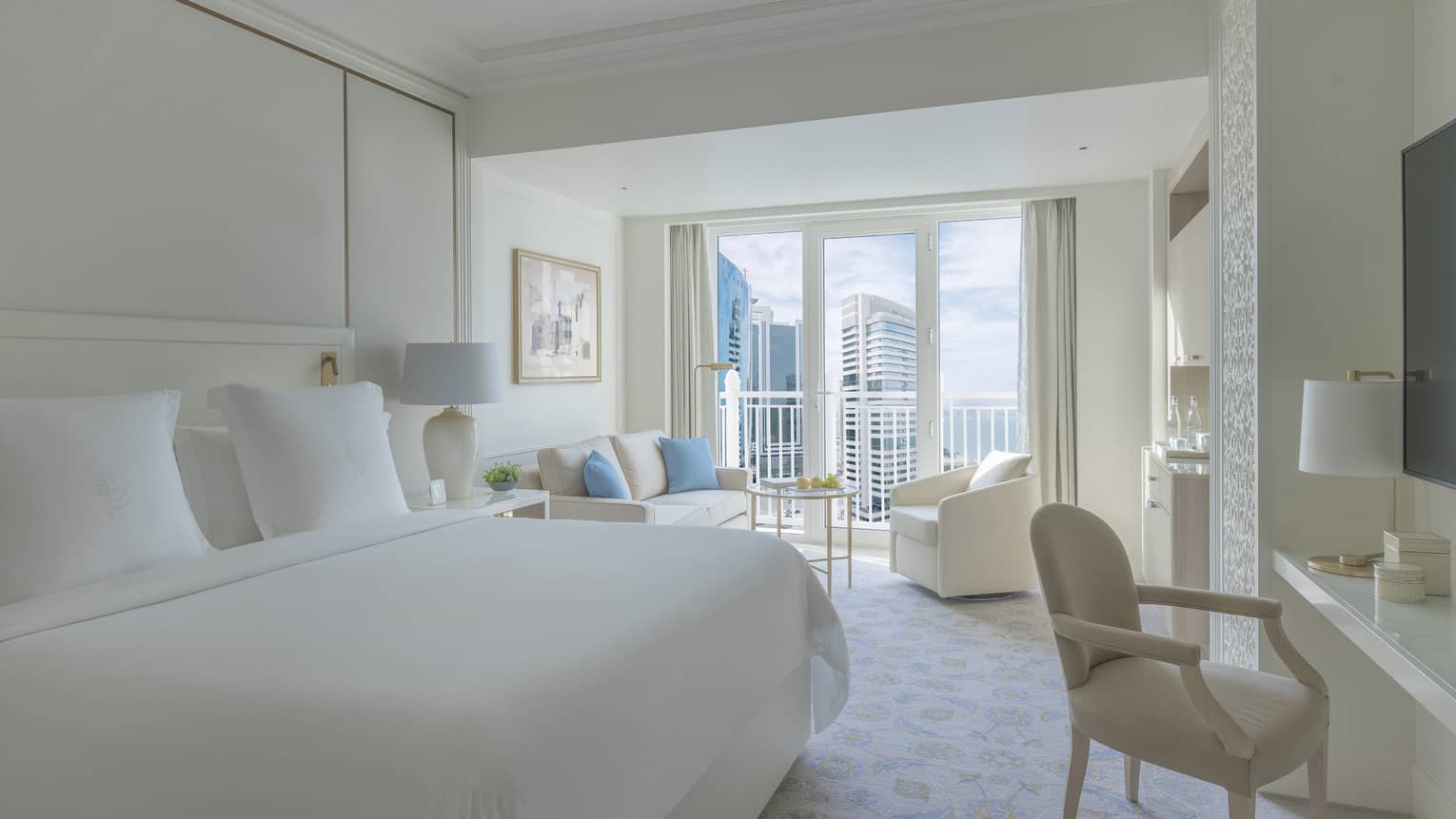 Bedroom with white bed, white walls and a white sofa and arm chair, with balcony offering city view