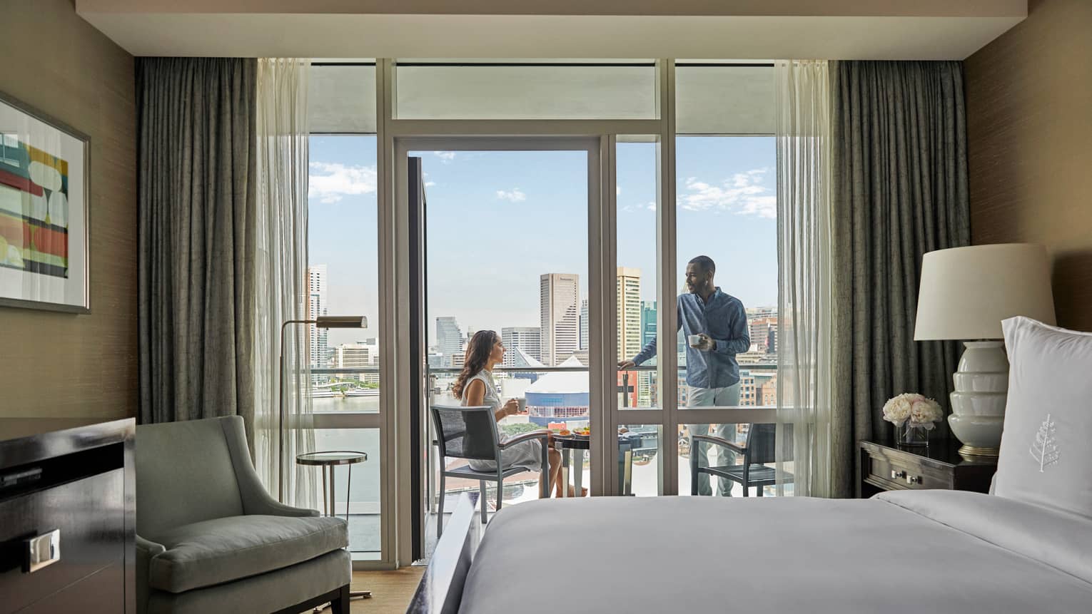 Hotel room with king bed, light grey arm chair, opening to outdoor balcony with two people talking