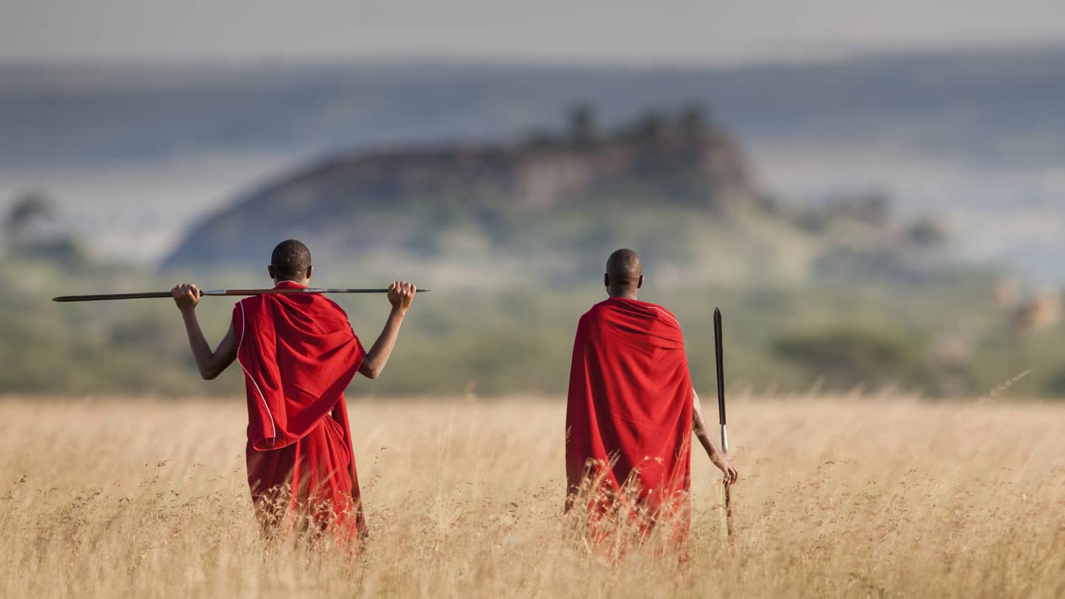 Two men in red indigenous clothing look out at a mountain on a savannah.