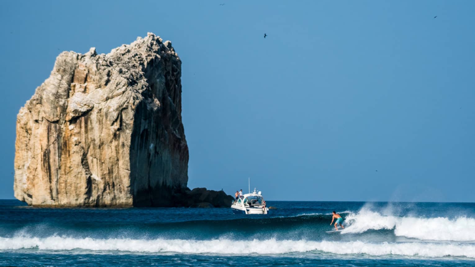A surfer rides a wave in the foreground, with a boat nearby and a large rock formation towering above the water in the background
