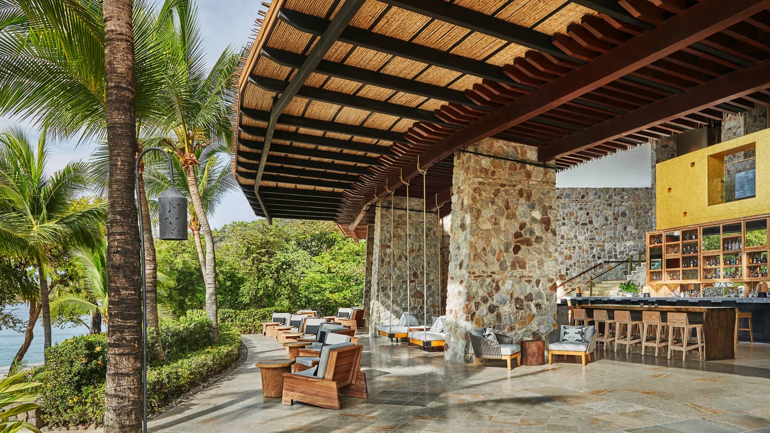 Anejo bar under large stone pillars, curved rattan roof over lounge chairs looking out at palms