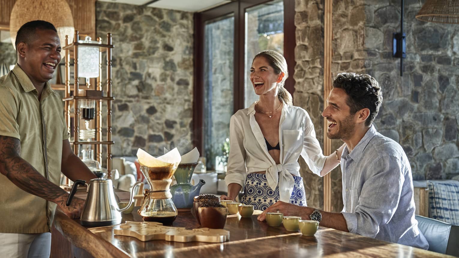 Carafes, cups of coffee and bowls of roasted coffee beans rest on a bar as two guests share a laugh with their bartender.