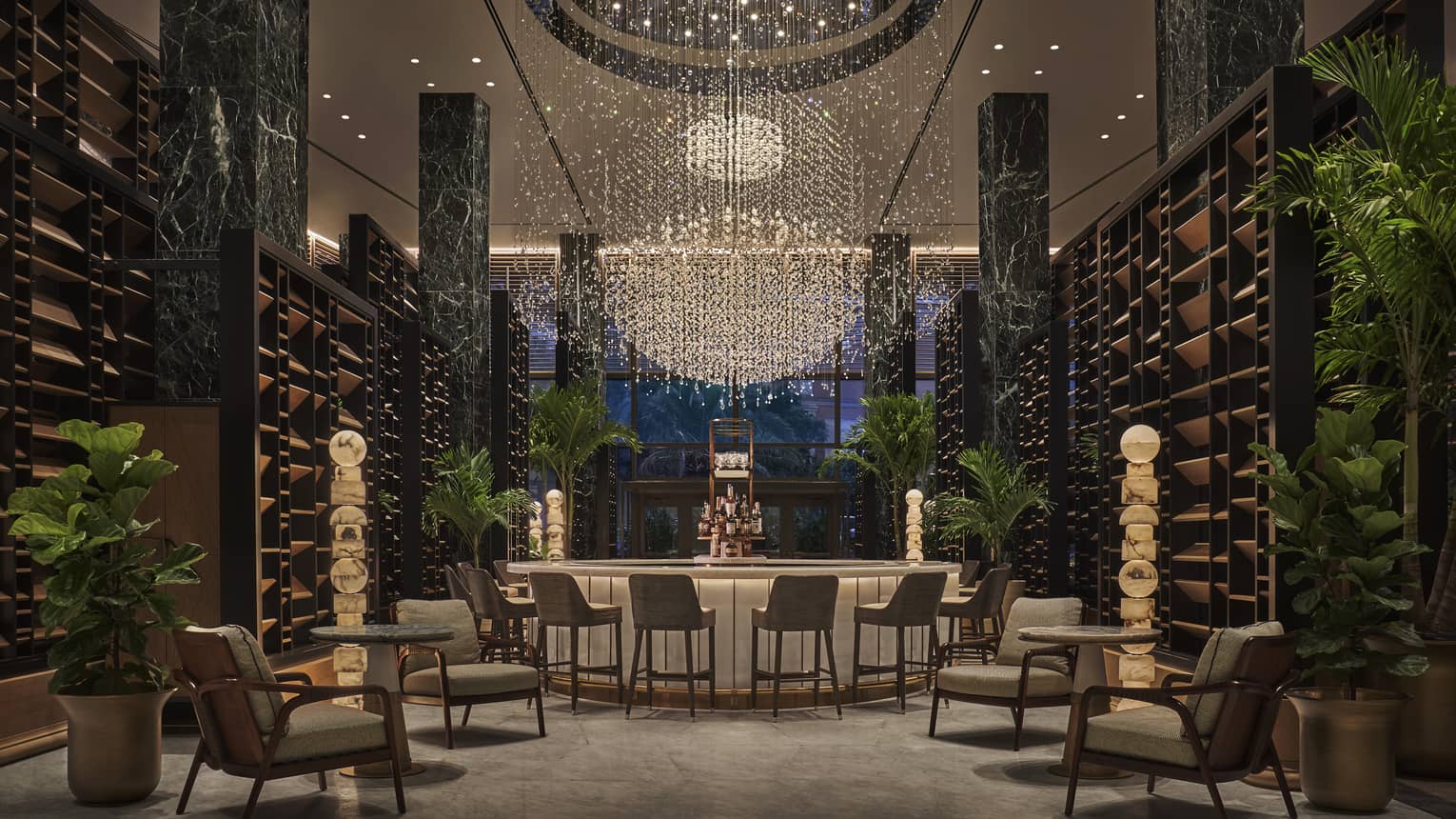 Chandelier Bar with six jade pillars flanked by wooden shelves and potted palms, glass-topped bar in the middle with six cocktail chairs, giant chandelier hanging overhead