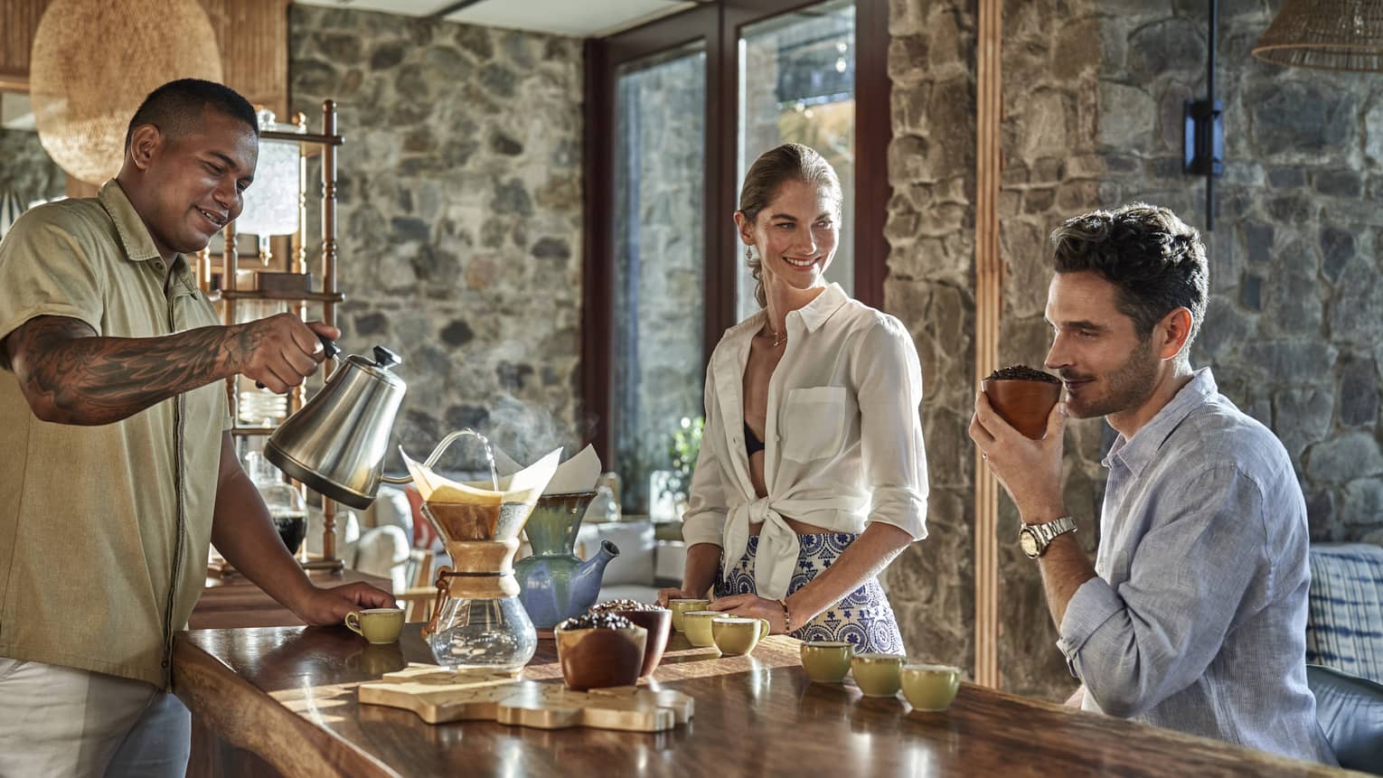 A man sits next to a wooden counter smelling a fresh cup of coffee, a woman stands next to him and another man stand on the opposite side pouring water into a chemex