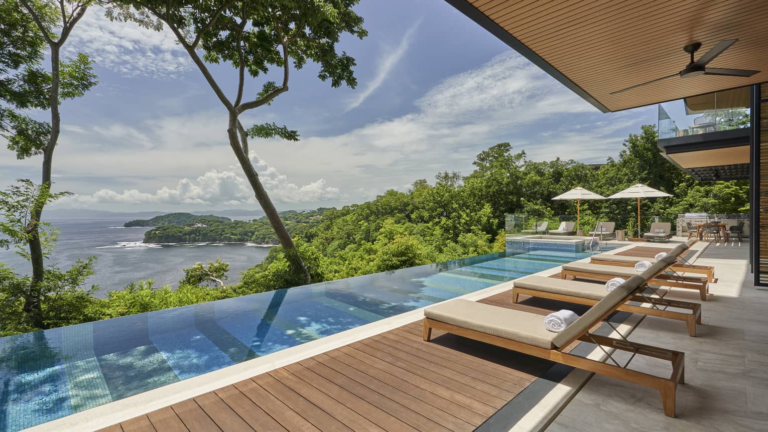 Narrow rectangular pool and wooden pool deck with cushioned lounge chairs, overlooking the sea