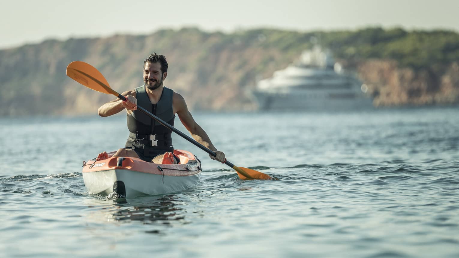 Man smiling while kayaking in sea, yacht and rocky coast in backdrop