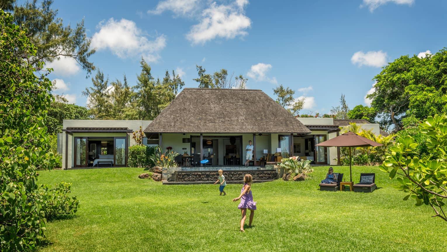 Young children play on green lawn in front of bungalow as dad watches from covered patio