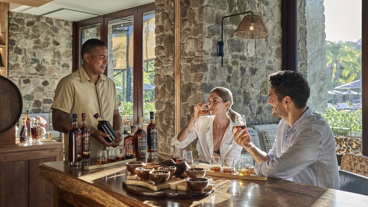 In a rustic bar with windows set between stone walls, a bartender looks on as two guests sample from a flight of eight rums.